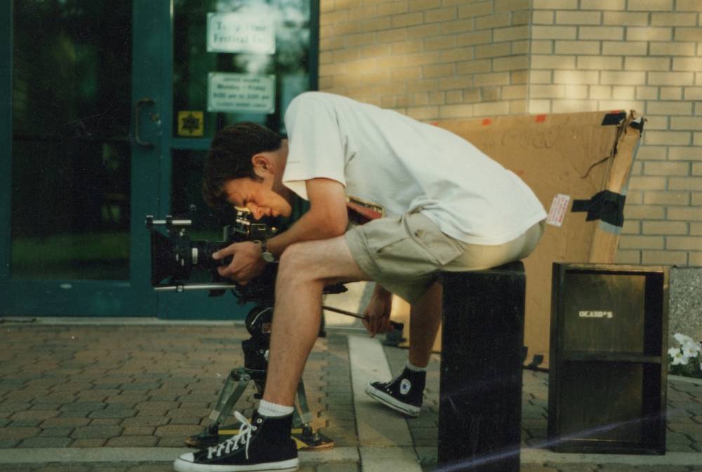 A camera operator looking through the lens of the camera while sitting on a black applebox